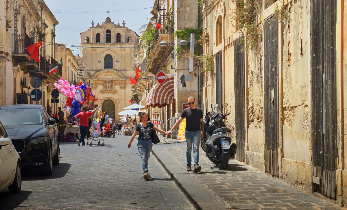 Narrow street in Noto with a baroque church facade at the far end and a few parked cars and scooters