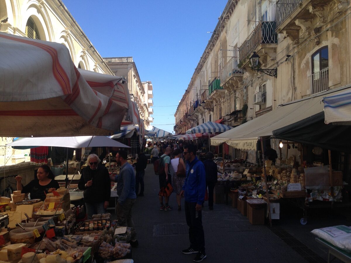 Market stalls and awnings along a narrow street in Ortigia, with shoppers browsing