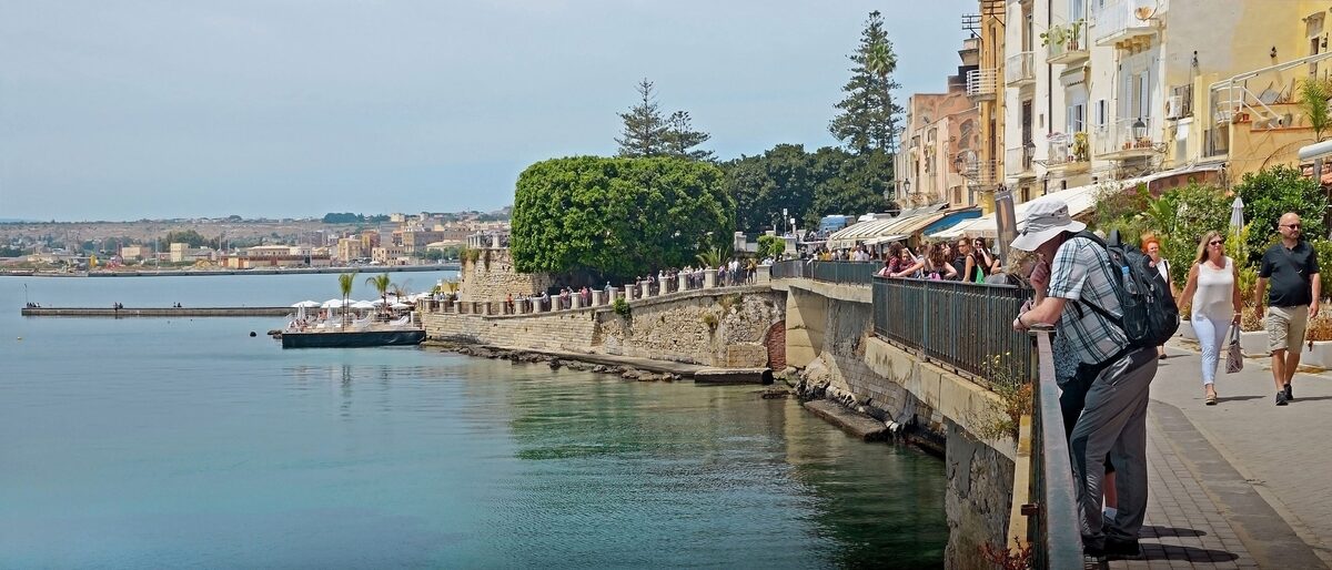 Seafront promenade of Ortigia above calm green water, with a stone wall, a lido platform and people at the railing
