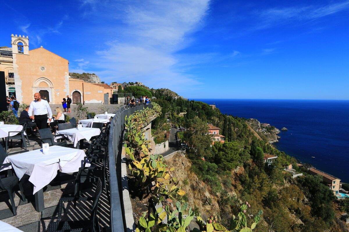 Taormina's clifftop terrace with a church facade and café tables, looking down over the wooded coast to the sea