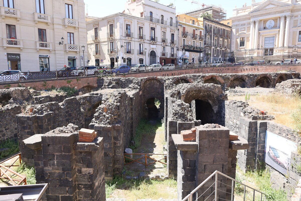 Black lava-stone arches and vaults of the Roman amphitheatre in Catania, with city buildings behind