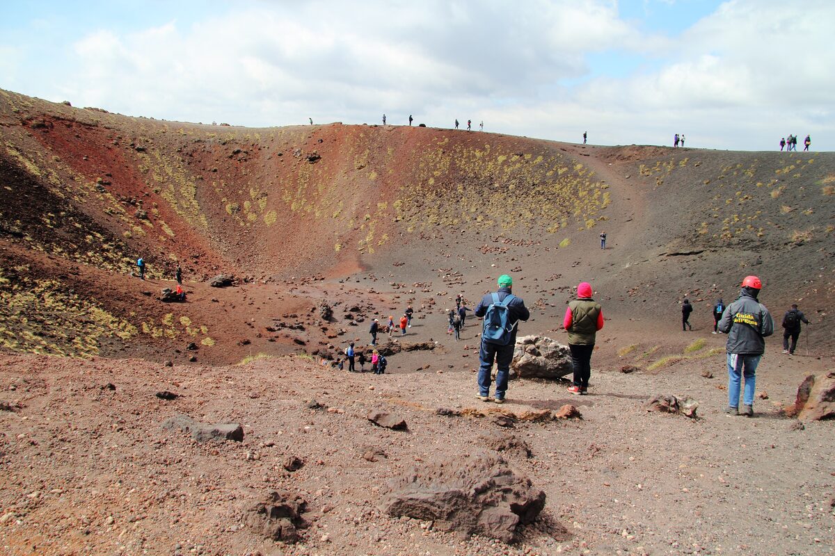 Red-rimmed volcanic crater on Mount Etna with hikers standing at the edge over a grey lava field
