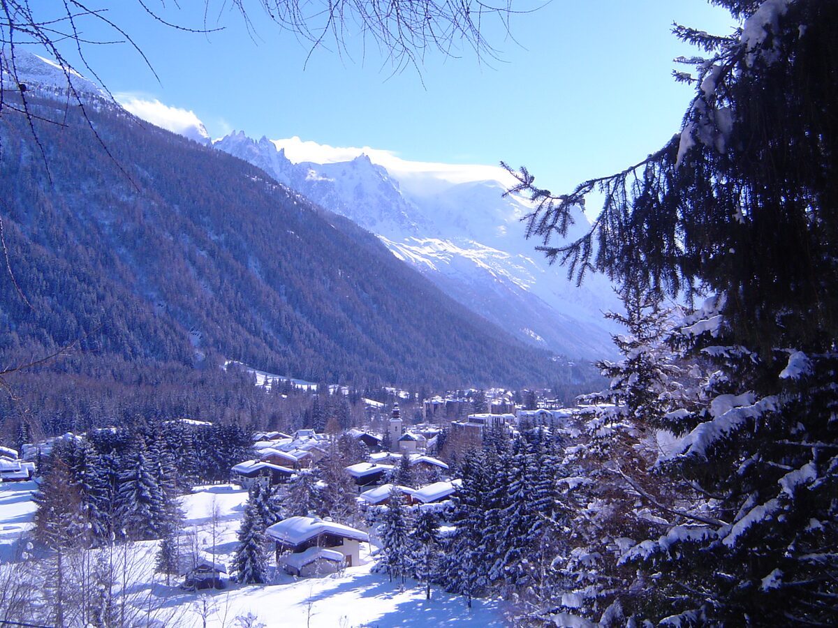 Snow-covered chalets and fir trees in the valley near Argentière on a sunny winter day, with the Mont Blanc massif at the end of the valley