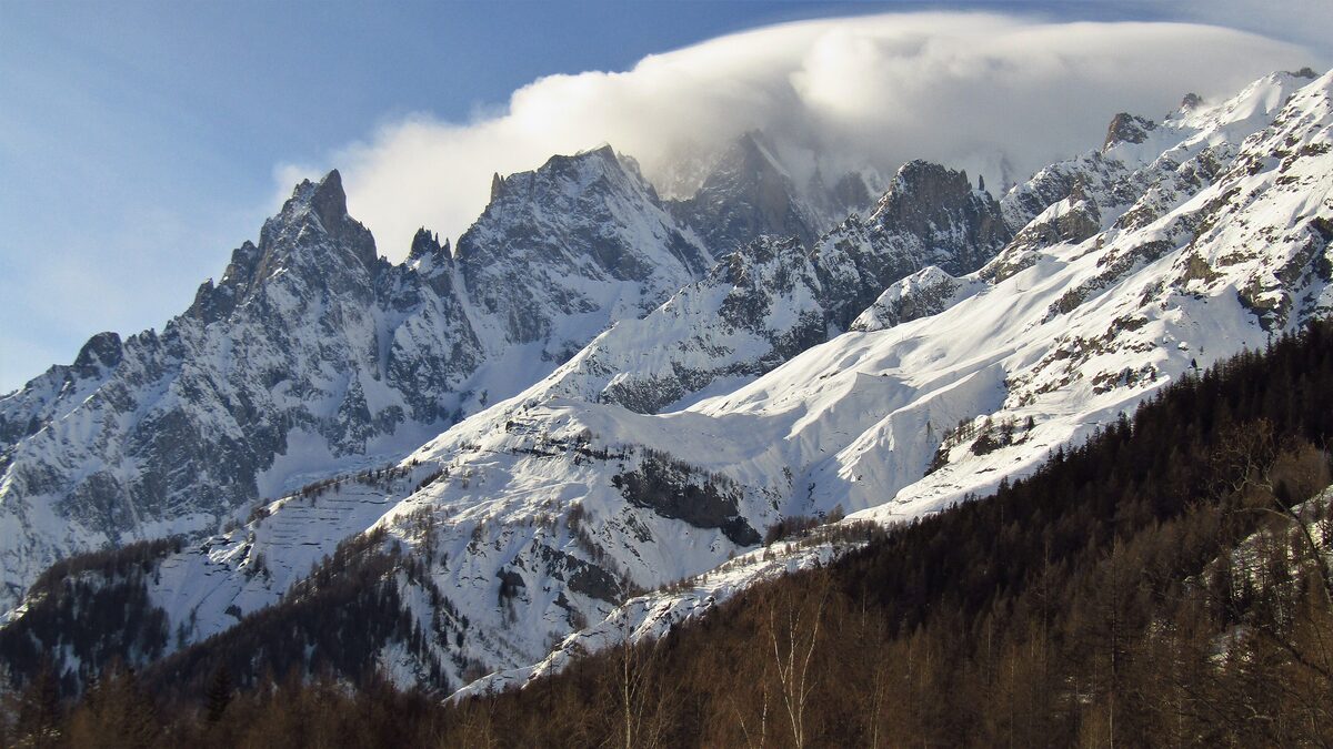 Snow-covered rock peaks of the Mont Blanc massif above the Val Ferret near Courmayeur, with a cloud cap on the summit and larch woods below