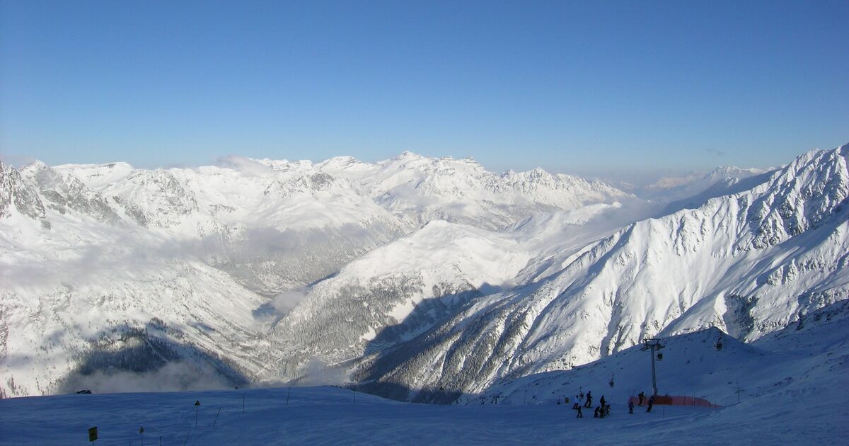 Skiers on a snowy piste above the Chamonix valley, with a lift pylon and snow-covered peaks beyond