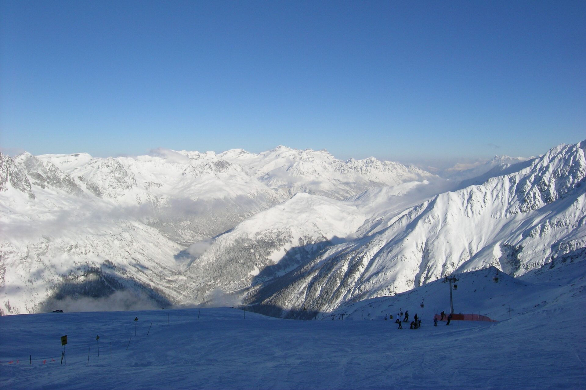 Skiers on a snowy piste above the Chamonix valley, with a lift pylon and snow-covered peaks beyond