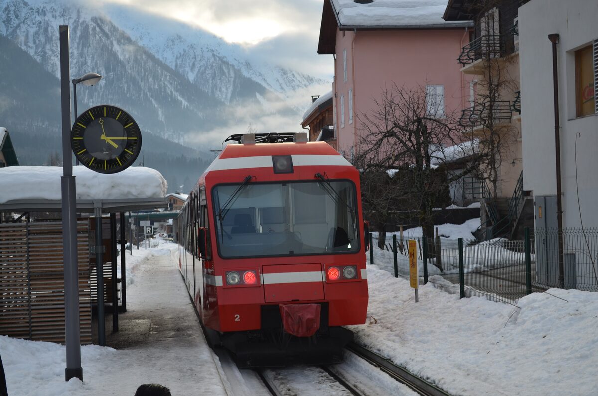 A red Mont-Blanc Express train arriving at a snowy station in the Chamonix valley, with a snow-covered roof, chalets and misty mountain slopes behind