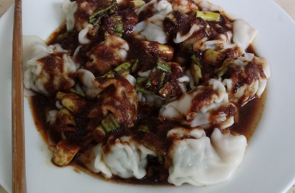 Wontons in red chili oil with chopped scallions on a white plate