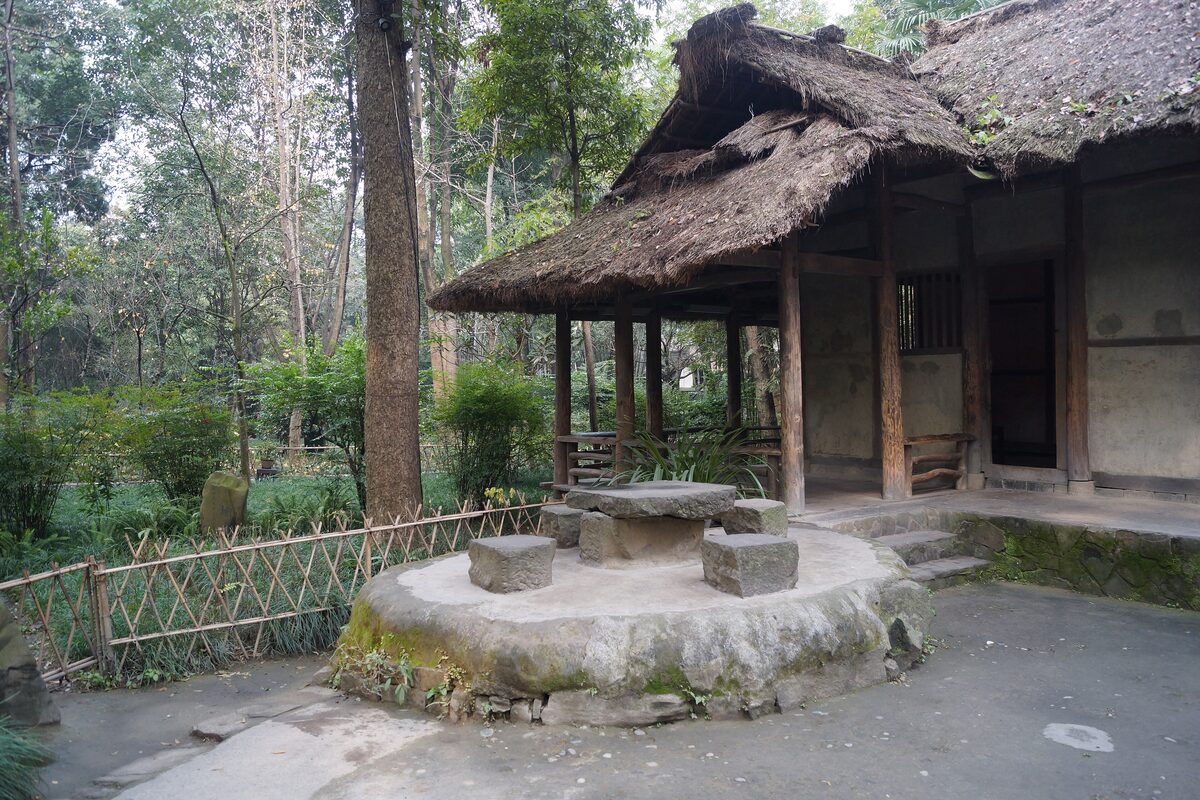 A thatched-roof hut and a round stone table among trees in the Du Fu Thatched Cottage grounds
