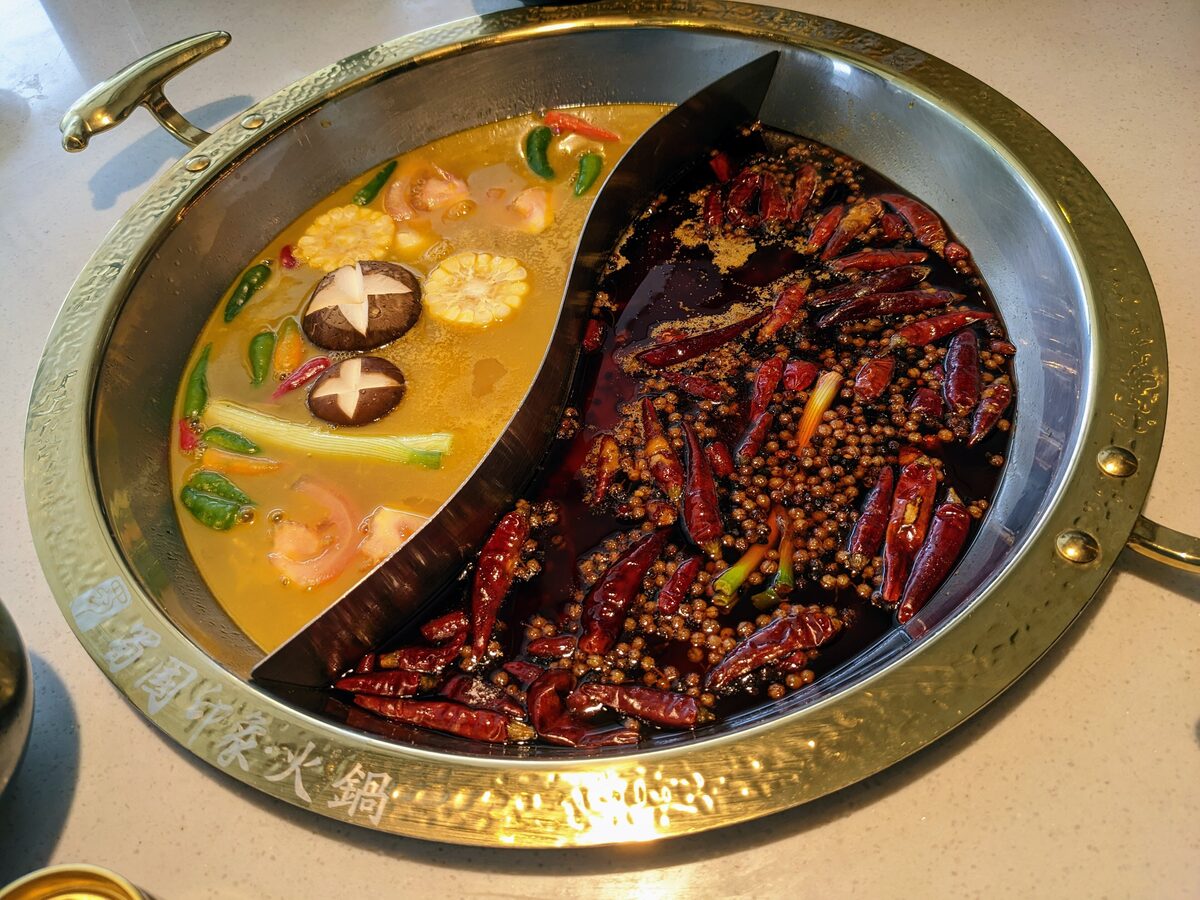A split Sichuan hotpot with a pale yellow mushroom-and-corn broth on one side and a red chili-and-peppercorn broth on the other