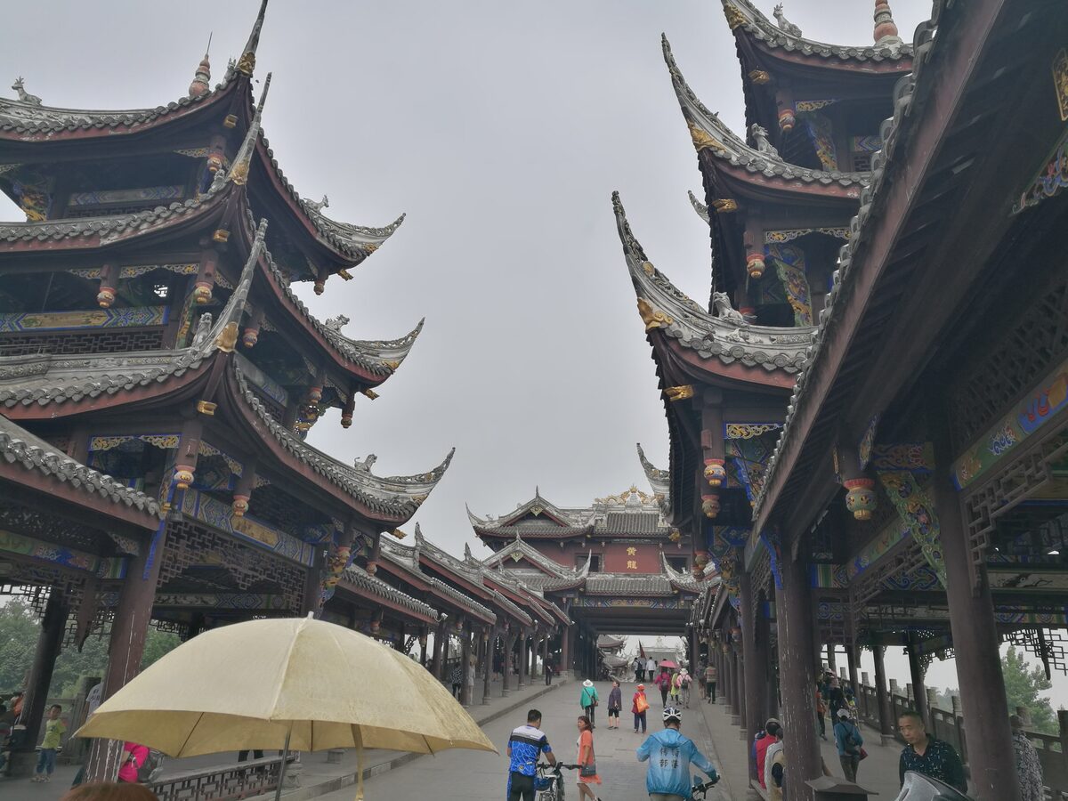 A covered wooden walkway with tiered roofs through Huanglongxi Ancient Town, with visitors under an umbrella