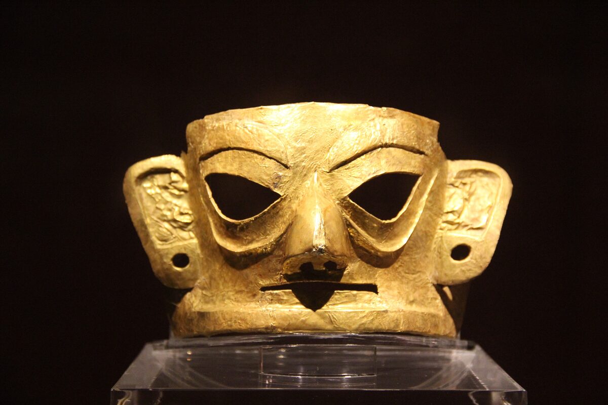 A gold mask from Jinsha with cut-out eyes and wide ears, lit against a black background in a museum case