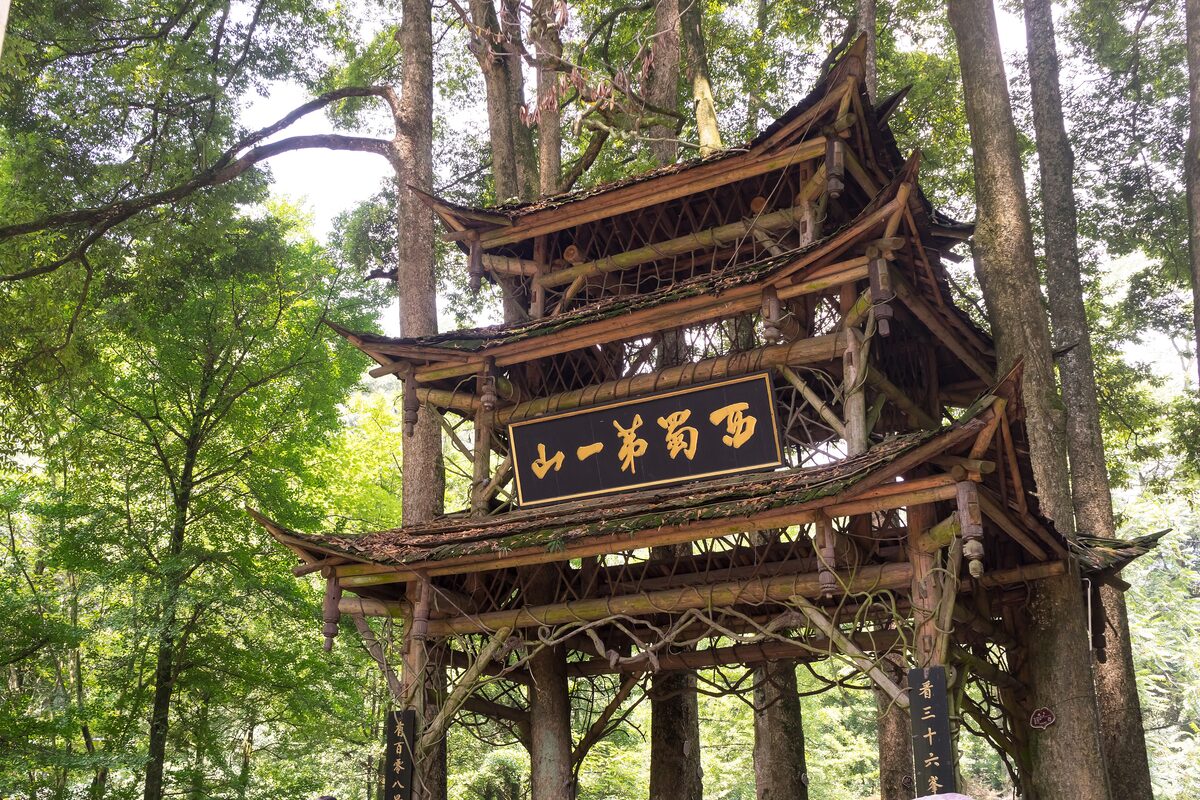 A wooden pavilion gate with a gilded plaque among tall trees on a Mount Qingcheng path