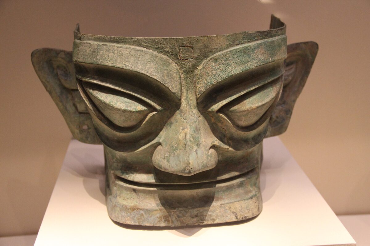 A bronze mask from Sanxingdui with bulging eyes and wide pointed ears, on a white stand in a museum case