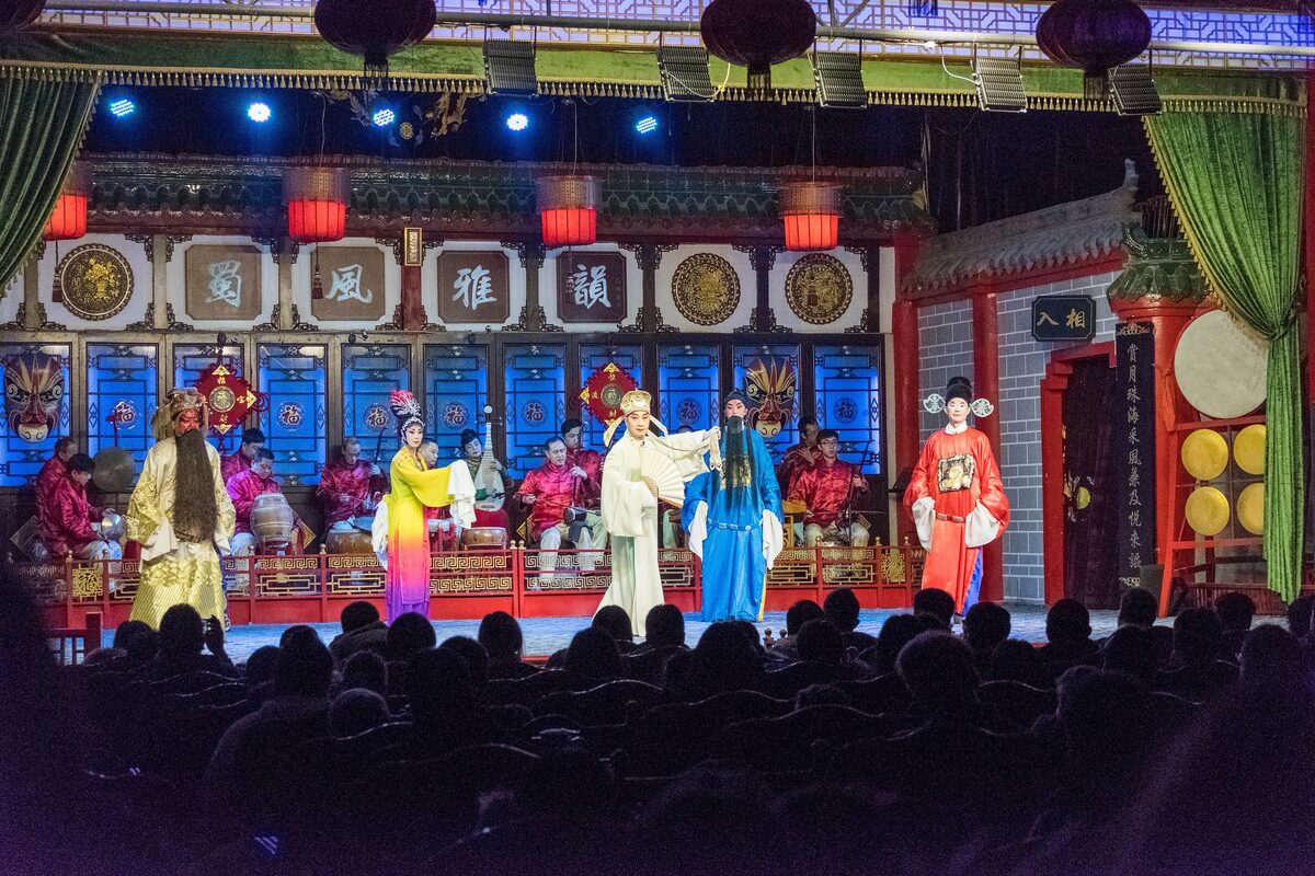 Sichuan opera performers in white, blue, yellow and red costumes on a lantern-lit stage at Shufeng Yayun, with an orchestra behind them and the audience in the foreground