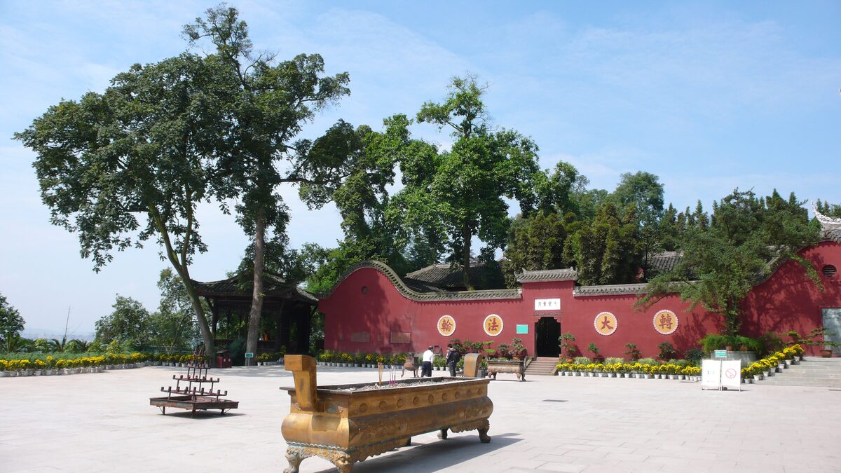 A bronze incense burner in the open forecourt of Wuyou Temple, with its red wall and old trees behind