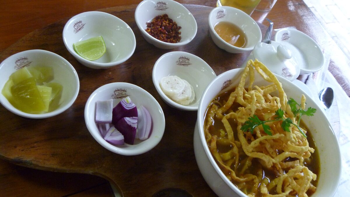 A bowl of khao soi with crispy noodles and small dishes of lime, chili, shallot and pickled greens