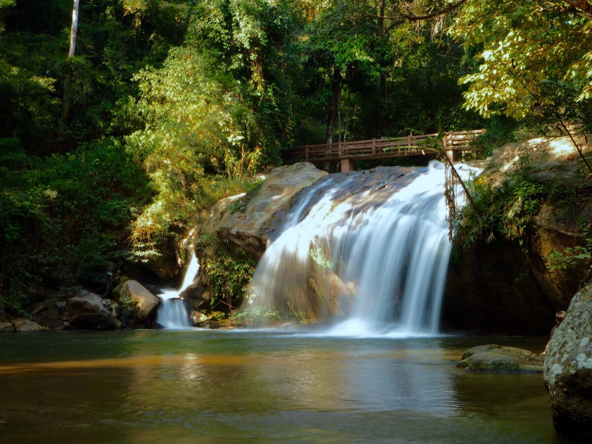 A tiered waterfall with a wooden footbridge above it and a calm pool below, in forest near Mae Sa