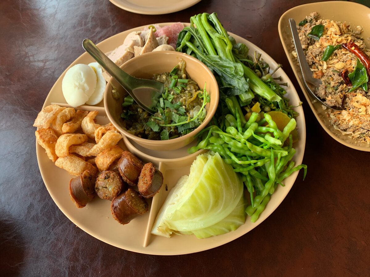 A small bowl of nam prik noom beside fried pork rinds, sausage and fresh vegetables