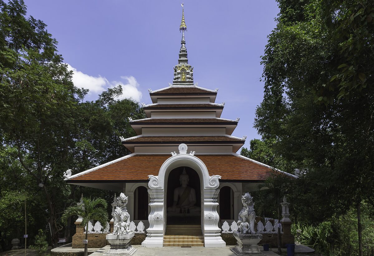 A white tiered shrine with guardian statues at its entrance, among trees at Wat Pha Lat
