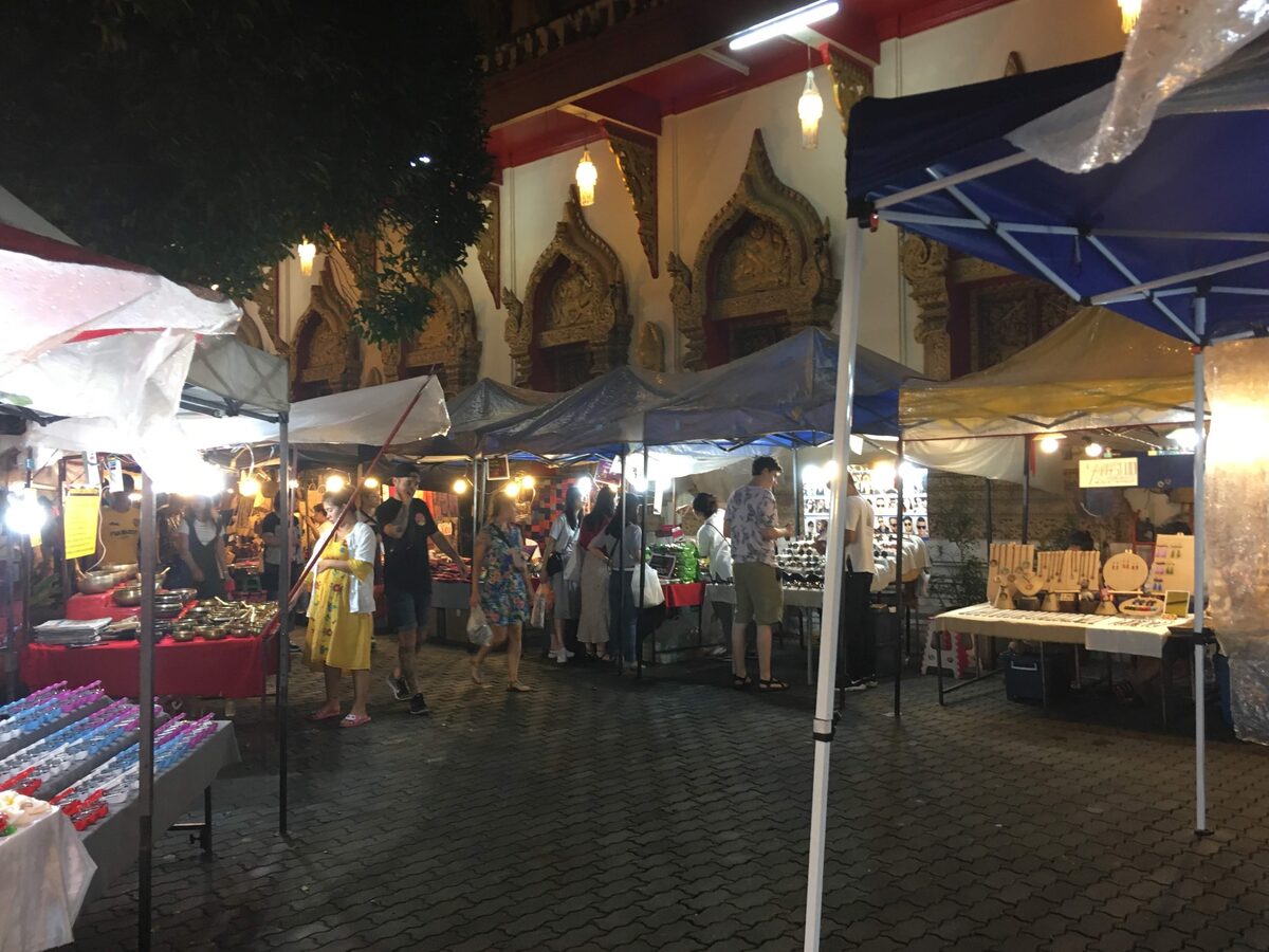 Lit market stalls with jewelry and crafts under tents beside a temple gate at the Sunday Walking Street at night