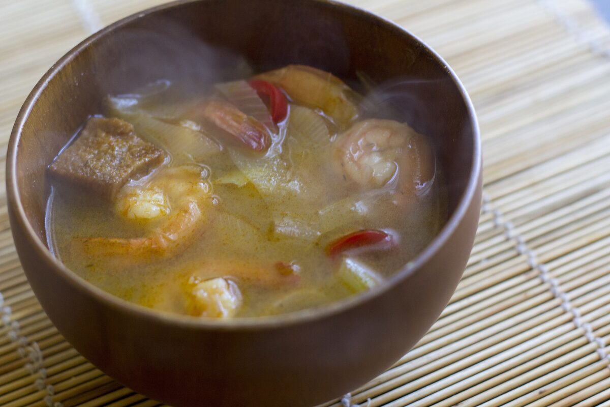 A steaming wooden bowl of creamy shrimp soup with chili and onion on a bamboo mat