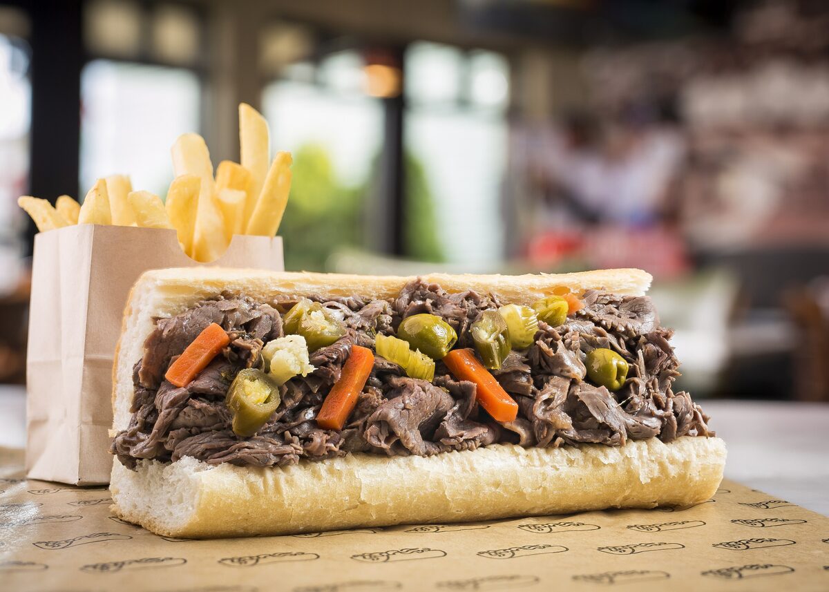 An Italian beef sandwich on a long roll topped with giardiniera and carrot slices, next to a bag of fries