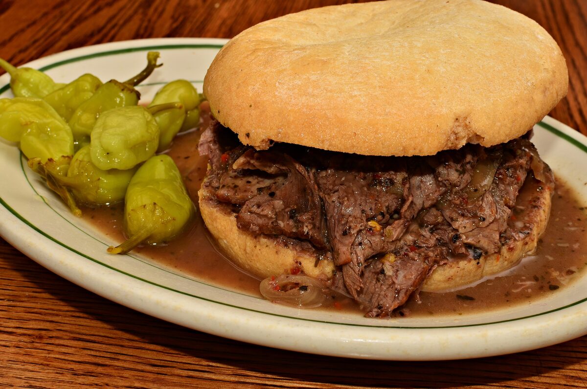 An Italian beef sandwich in its gravy on a plate, with pickled peppers on the side