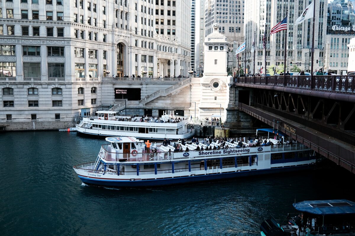 Two sightseeing boats on the Chicago River beside a bridge, with white terra-cotta buildings on the bank
