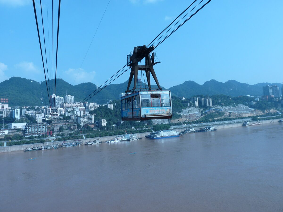 A Yangtze River Cableway car on its cables above the brown river under a blue sky, with green hills and the far bank behind