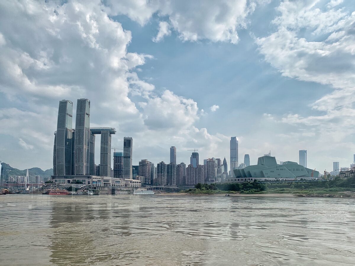 The Raffles City towers at the tip of the Chongqing peninsula seen across the brown Yangtze under a blue sky with clouds