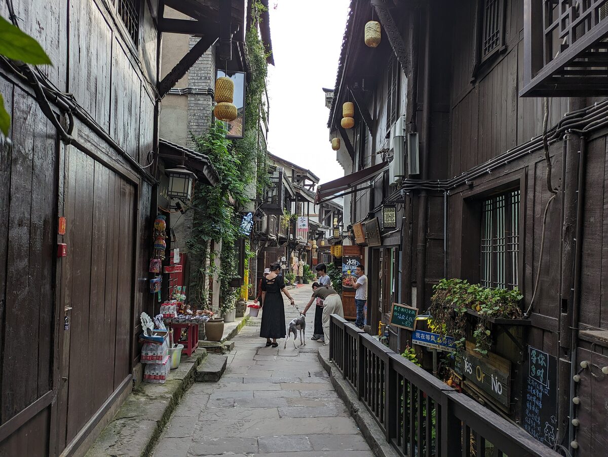 A narrow stone-paved lane in Ciqikou old town between dark timber houses, with hanging lanterns and a few walkers