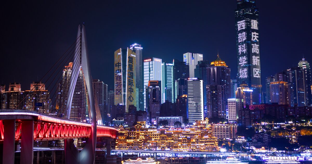 The Chongqing skyline at night across the Jialing River, with Qiansimen Bridge in red and the lit Hongyadong stilt houses at the waterline