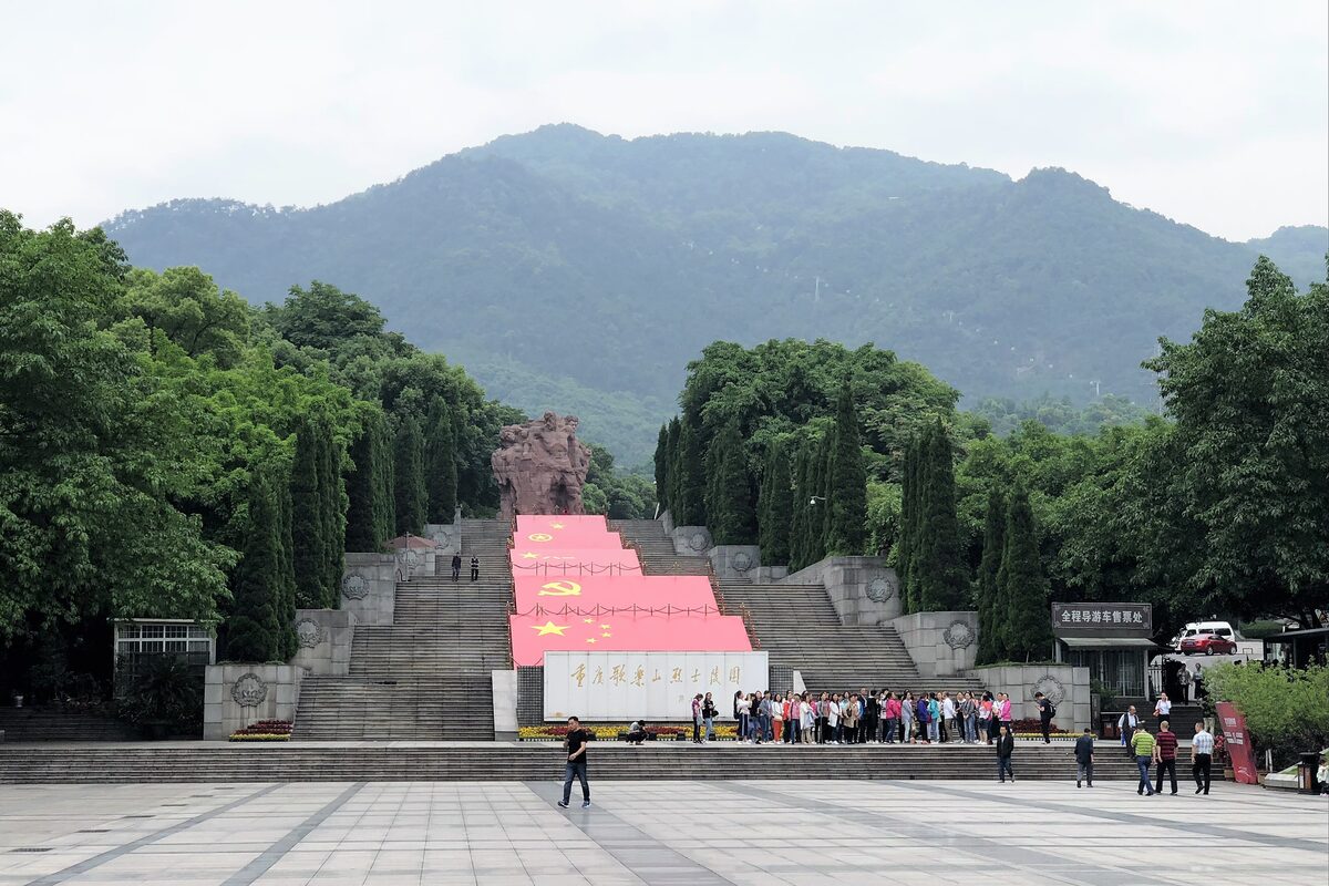 The wide stone stairway of the Gele Mountain martyrs' cemetery, draped with red flags and climbing toward the wooded hill, with a tour group on the plaza