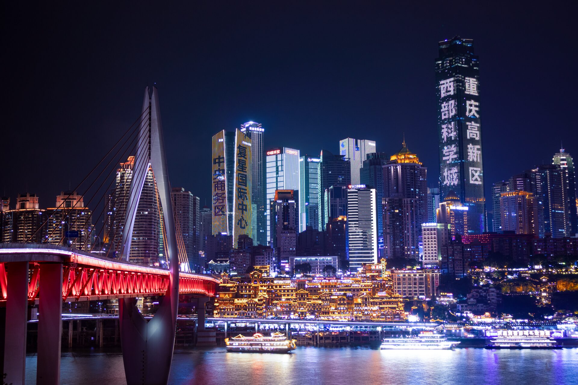 The Chongqing skyline at night across the Jialing River, with Qiansimen Bridge in red and the lit Hongyadong stilt houses at the waterline