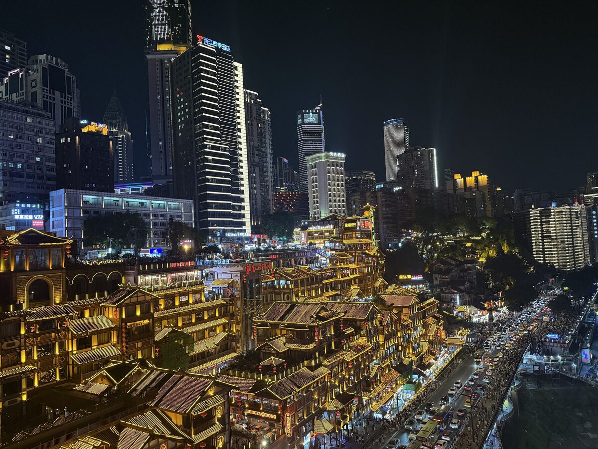 Hongyadong's tiers of stilt-style houses glowing gold at night, with a busy road and skyscrapers behind