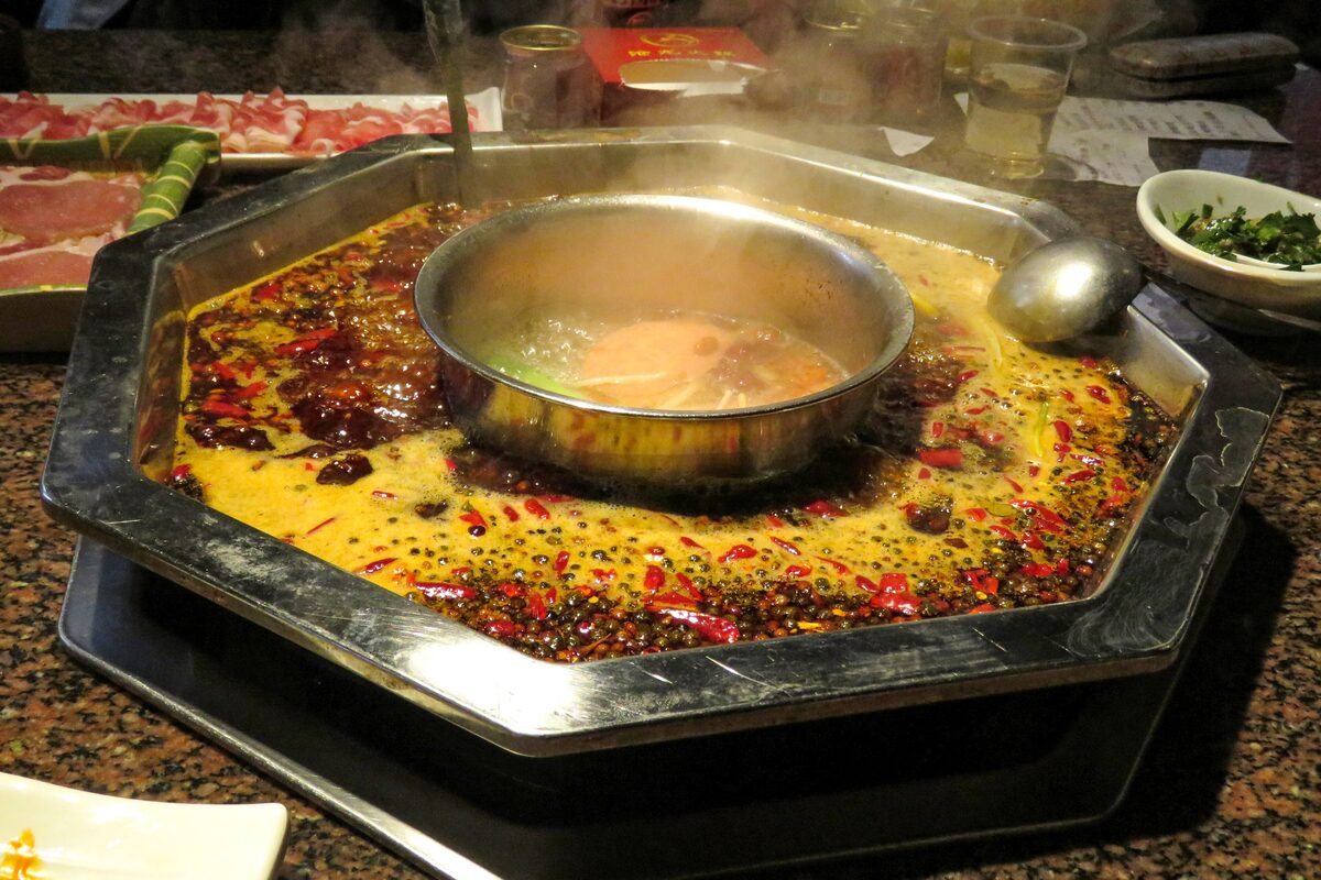 A steaming octagonal hot pot in Chongqing, its broth thick with dried chilies and Sichuan peppercorns around a small central bowl, with plates of sliced meat behind