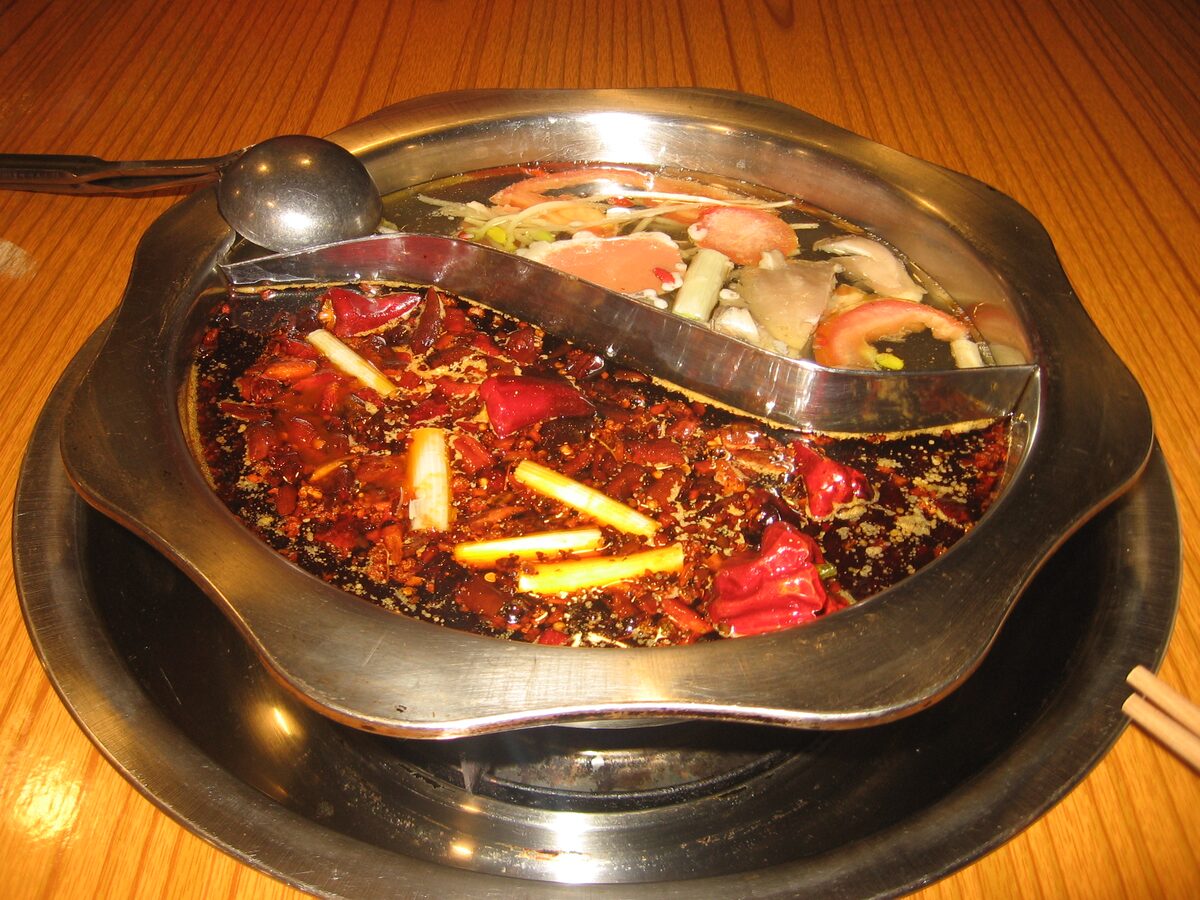 A two-flavor hot pot on a wooden table, one half dark red with dried chilies and scallion stalks, the other a clear broth with sliced vegetables and meat