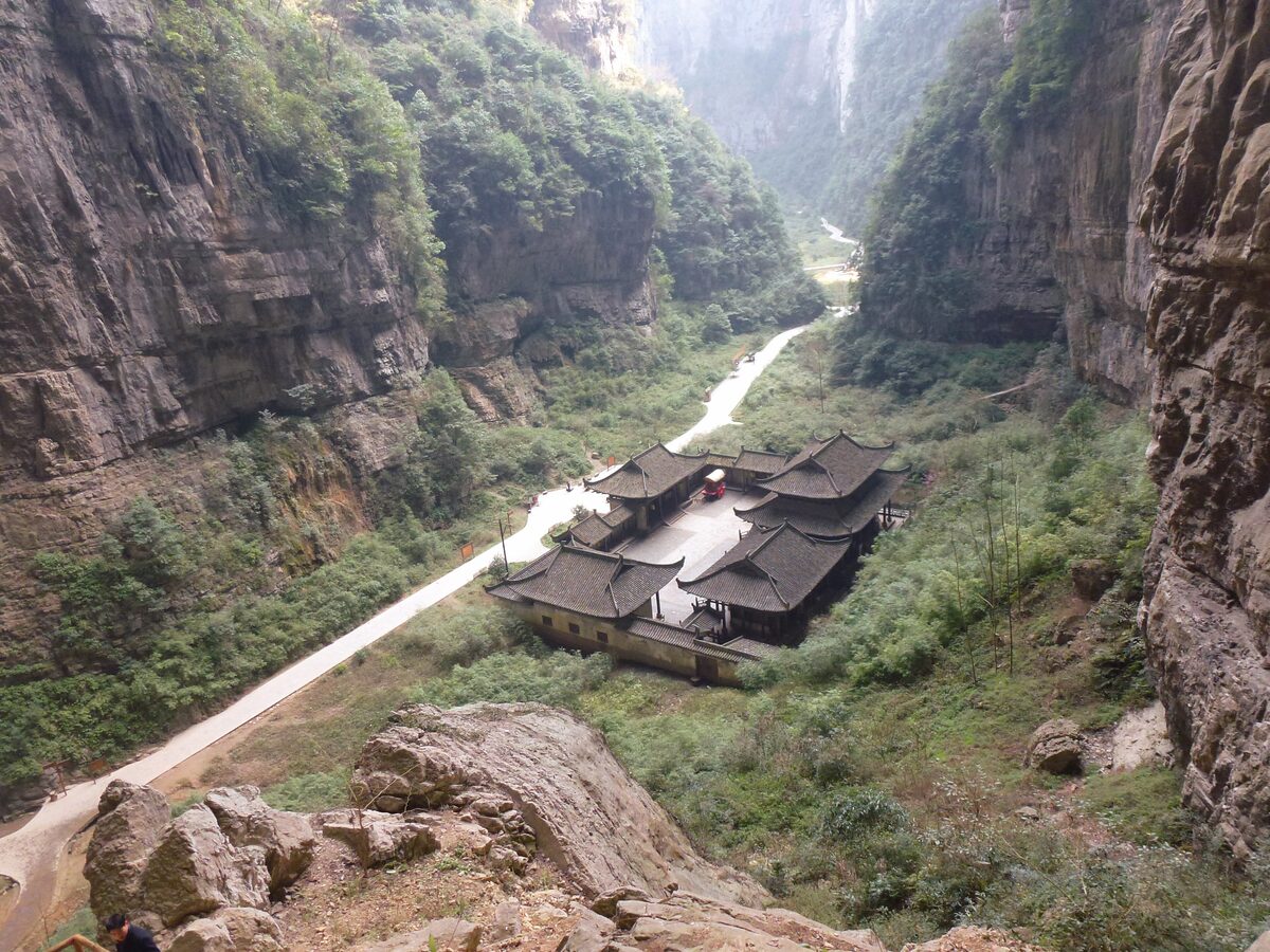 A courtyard of tile-roofed timber halls on the floor of a deep Wulong sinkhole gorge, with a path winding between steep cliffs
