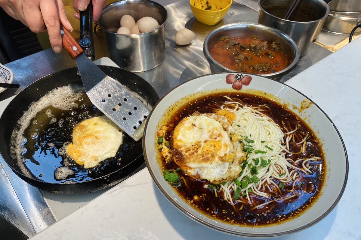 A bowl of xiaomian with a fried egg, scallions and dark seasoned broth at a noodle counter, a second egg frying in a pan beside it