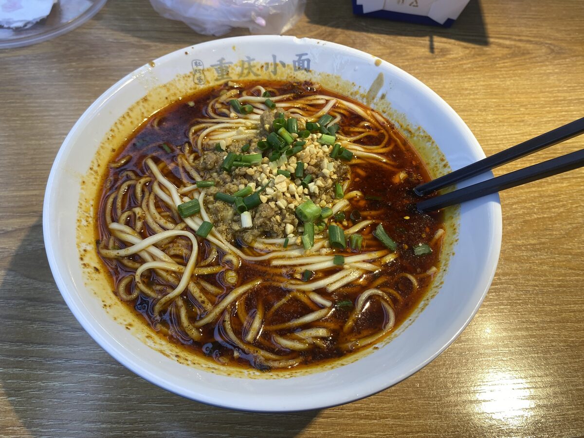 A bowl of Chongqing xiaomian in dark chili-oil broth topped with crushed peanuts and scallions, with chopsticks