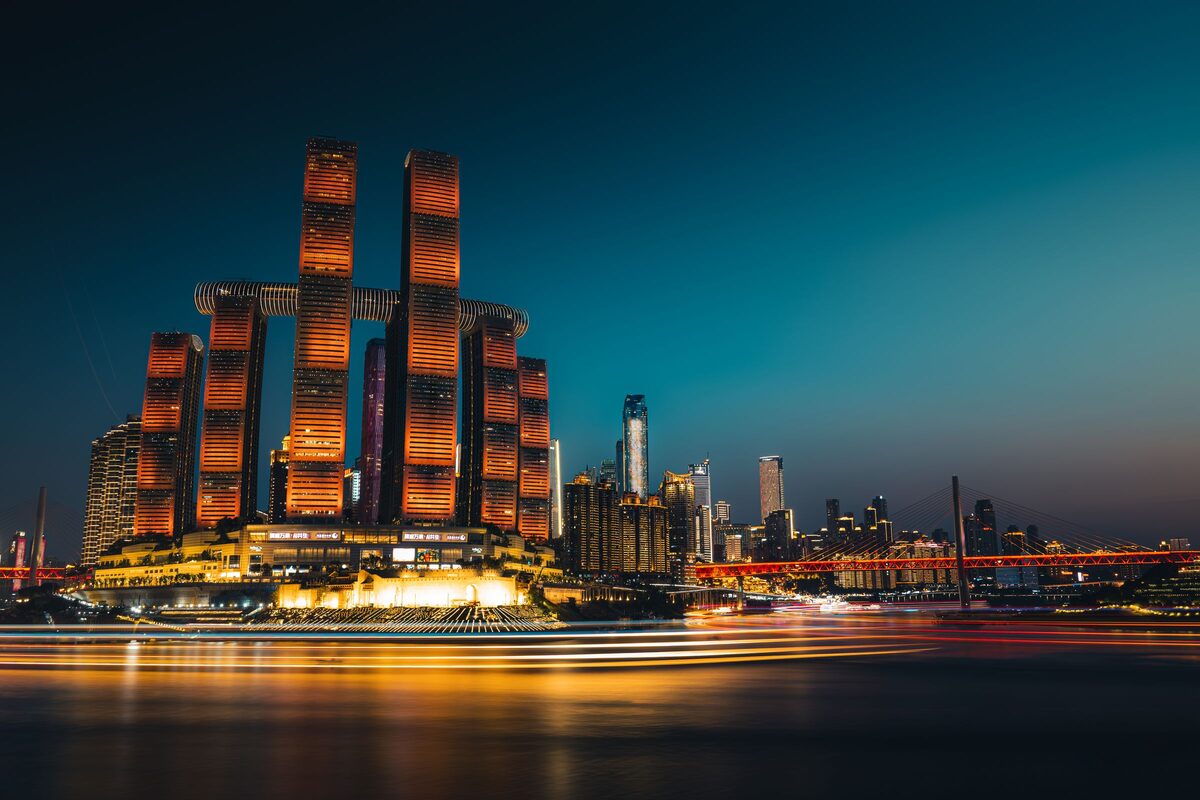 The lit Raffles City towers on the Chongqing peninsula across the Yangtze at blue hour, with a red bridge to the right and light trails on the river