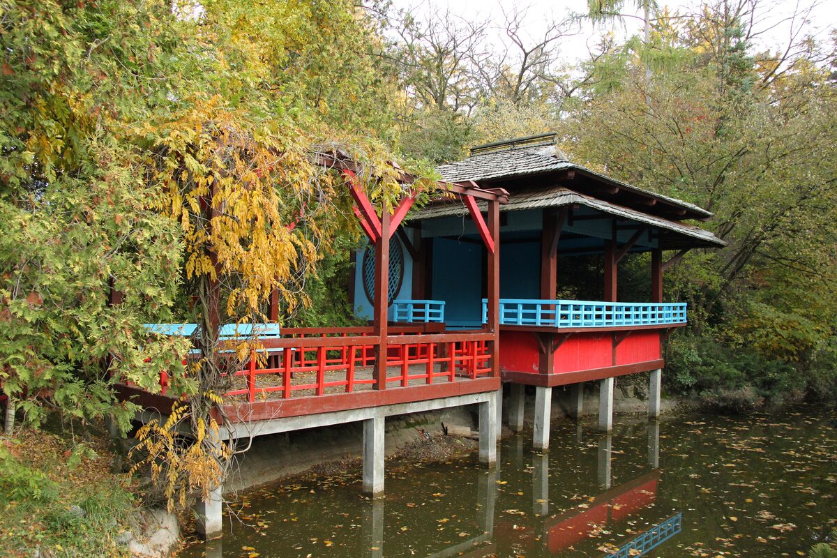 The red and blue Japanese tea house on stilts over the pond in the Japanese Garden of Cluj's Botanical Garden, framed by autumn foliage