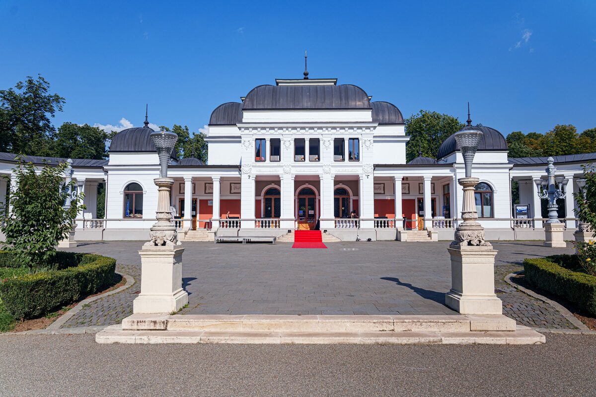 The white Casino pavilion in Cluj's Central Park under a blue sky, seen across a paved forecourt with ornate lamp posts