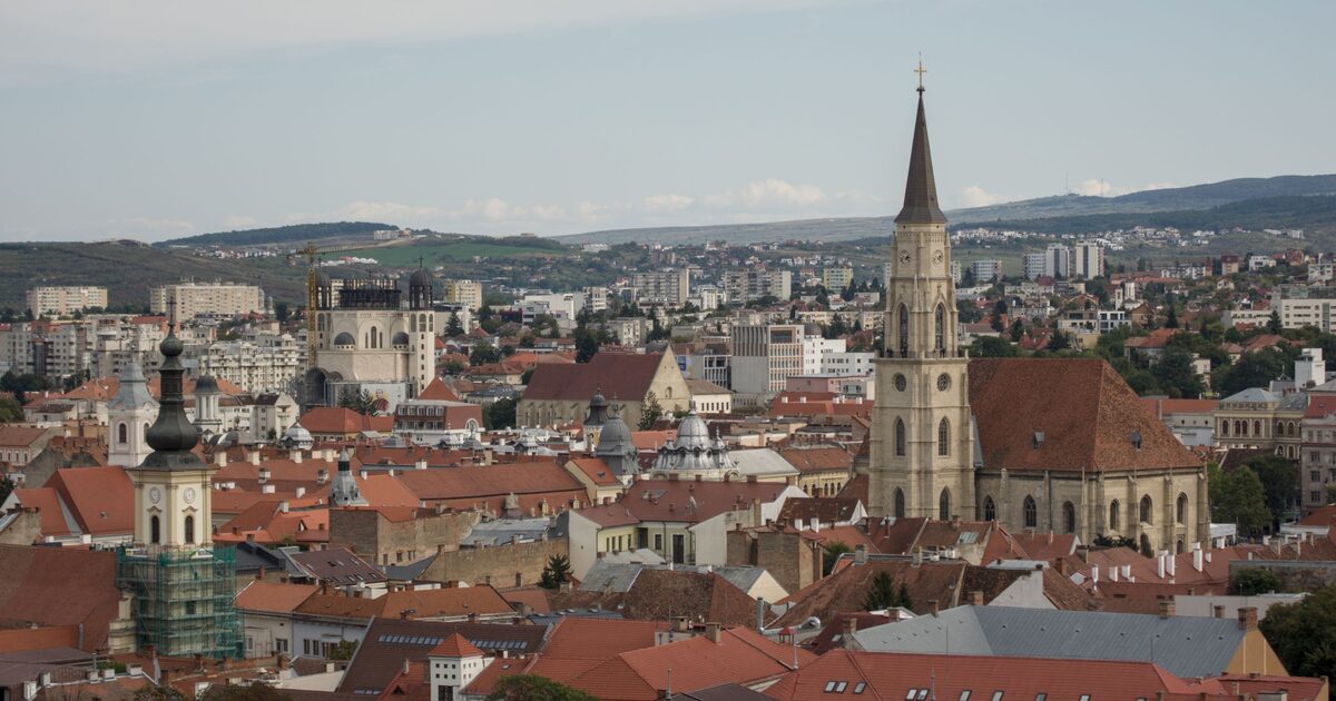 Elevated view over Cluj-Napoca's red-tiled rooftops, with the tall Gothic spire of St. Michael's Church and green hills on the horizon