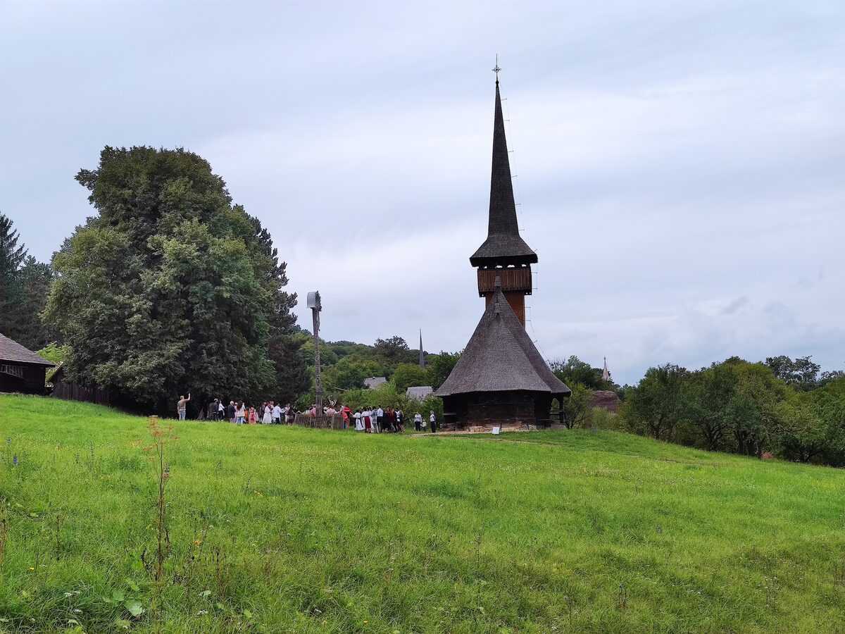 The dark wooden church from Cizer with its tall, slender shingled spire, standing in a green meadow at the Romulus Vuia Ethnographic Park
