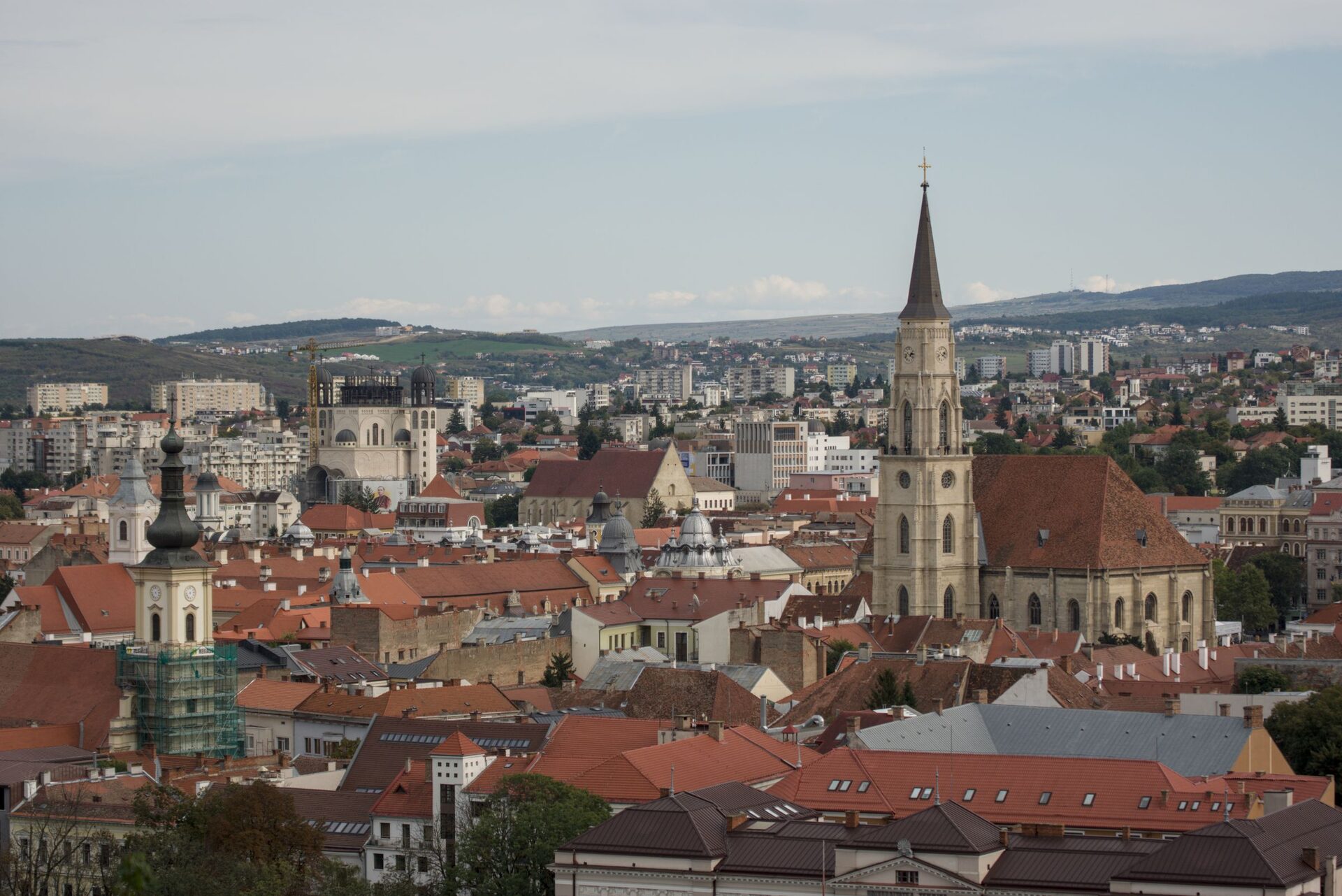 Elevated view over Cluj-Napoca's red-tiled rooftops, with the tall Gothic spire of St. Michael's Church and green hills on the horizon