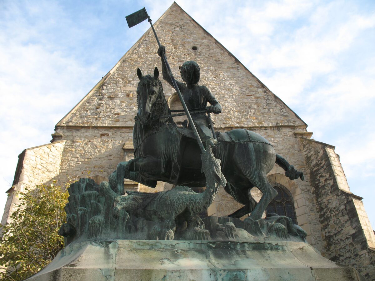 The bronze statue of St. George slaying the dragon, seen from below in front of the stone gable of the Reformed Church on Farkas Street