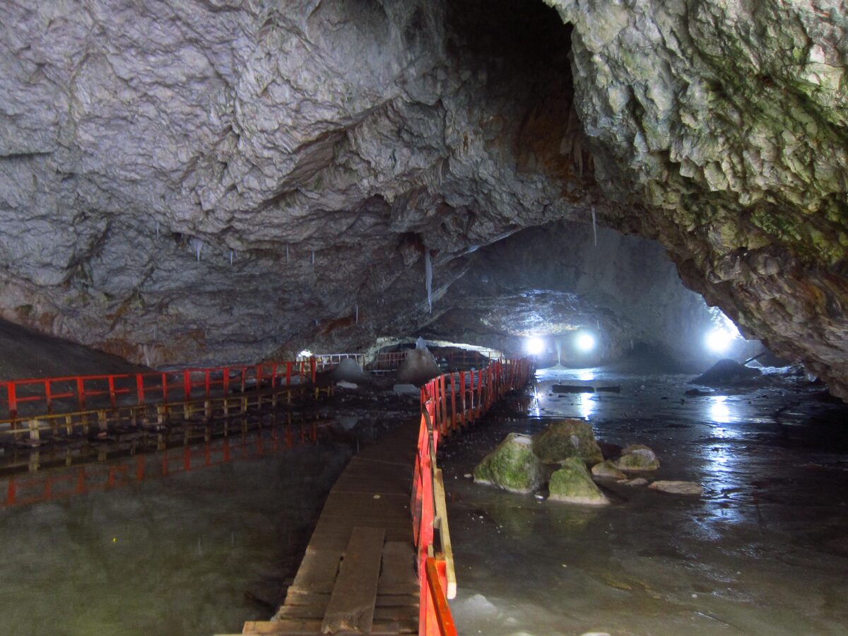 A wooden walkway with red railings crossing the ice floor of Scărișoara Ice Cave beneath a huge rock vault