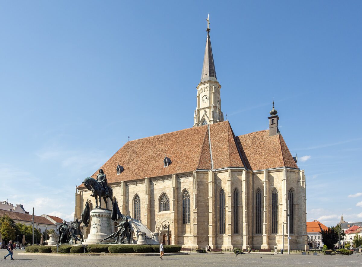St. Michael's Church on Piața Unirii with its tall neo-Gothic tower and the Matthias Corvinus equestrian monument in front, under a clear blue sky
